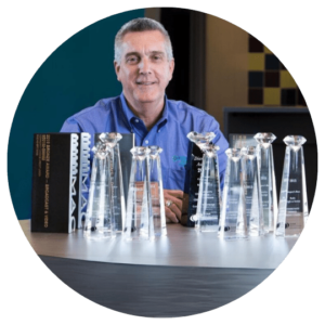 Alan Bergstrom smiles in a blue shirt sitting behind a desk, proudly displaying a collection of CUNA diamond awards and various AV awards.